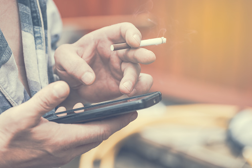 man-hand-holding-smoking-cigarette-and-using-mobile-smart-phone-toned-picture-id825477626